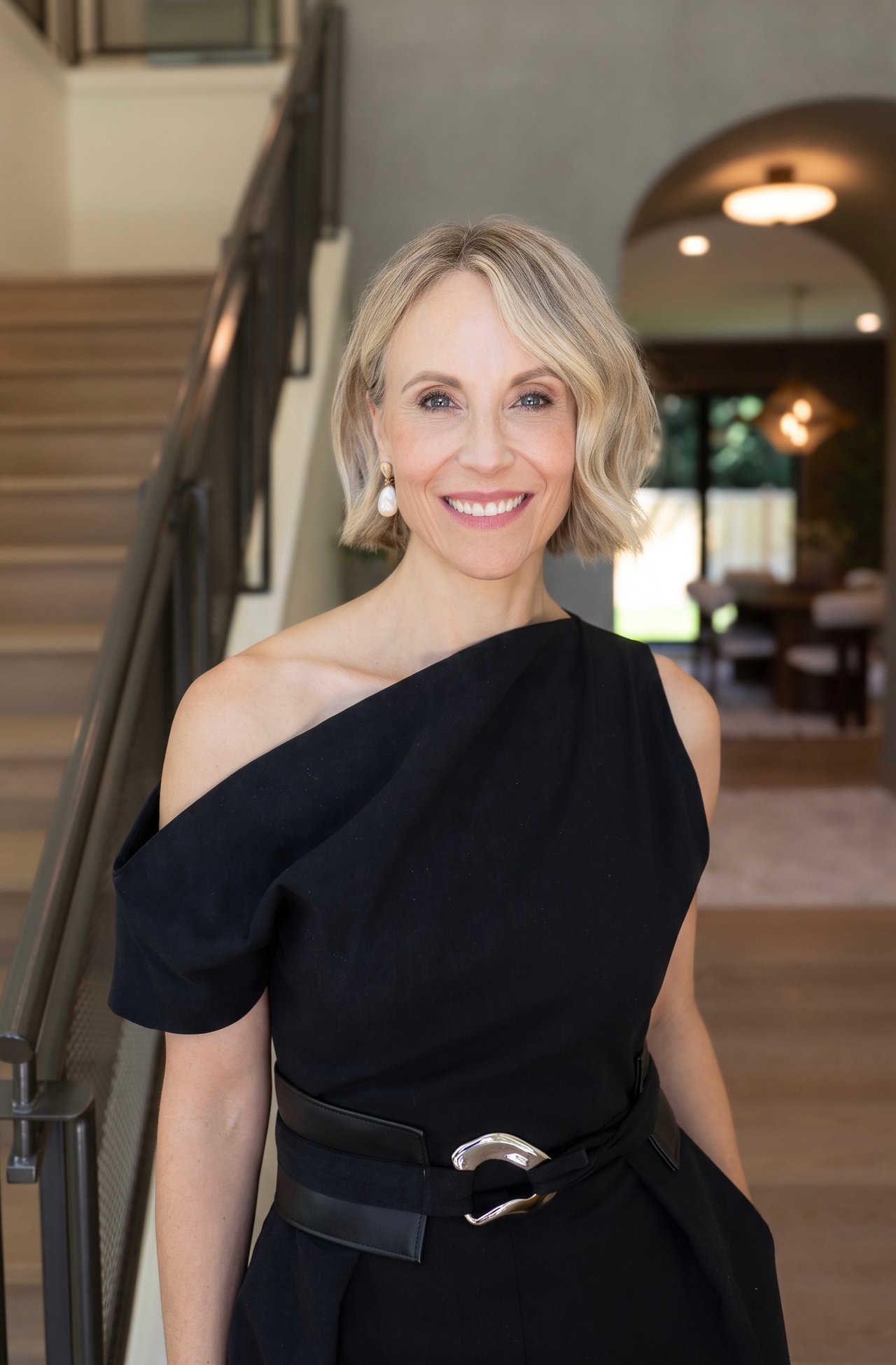 Ashley Rey smiles in a black one-shoulder dress beside a staircase.