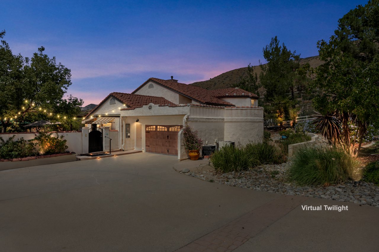Stucco home with terracotta roof and wooden garage door, lit at dusk beside a landscaped yard.
