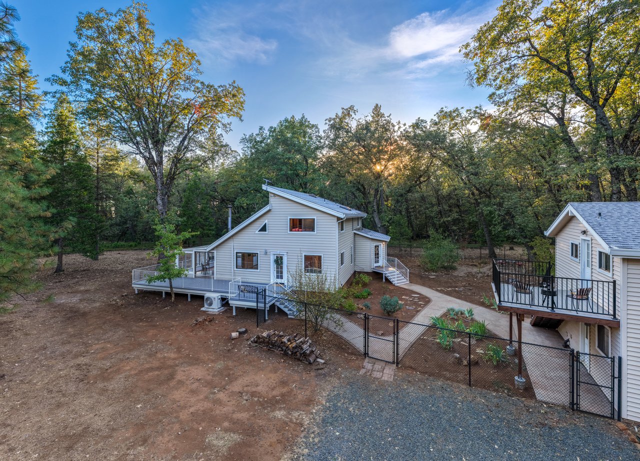 Light-colored home with a broad deck and fenced yard among mature trees at golden hour.