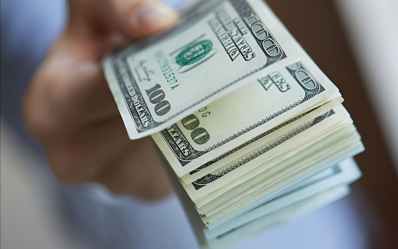 A hand holds a thick stack of hundred-dollar bills, showing the top bill and layered edges.