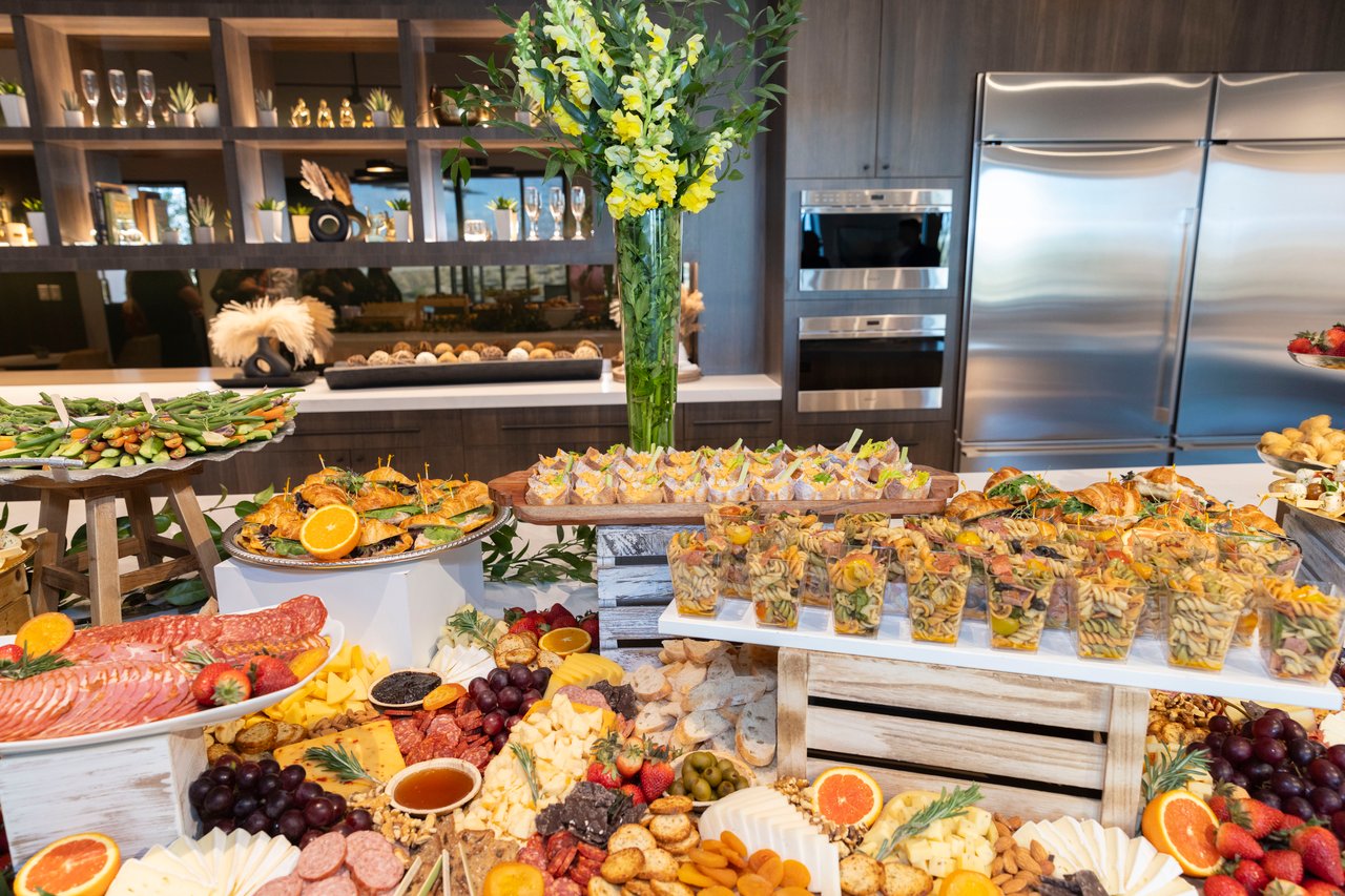 Open-house buffet with charcuterie, cheeses, fruit, pasta salad, and appetizers beneath yellow flowers.