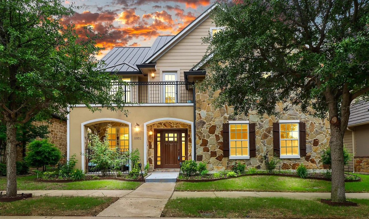 Two-story stone-and-siding house with a metal roof and warmly lit windows at dusk.