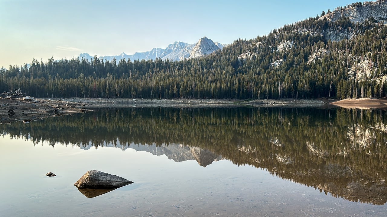 Mountain lake reflecting evergreen forest and rugged peaks, with rocks in the shallow foreground.