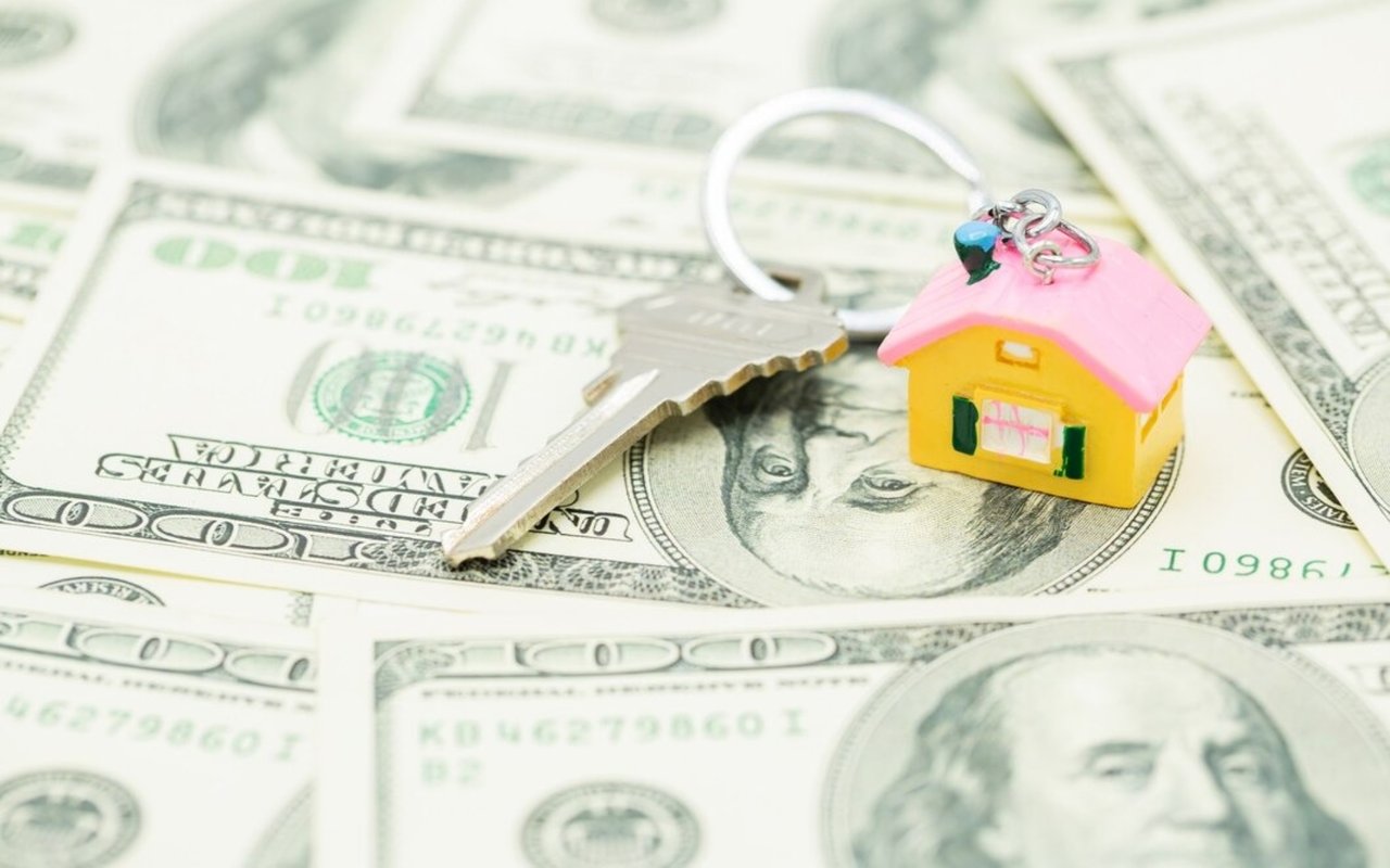 A miniature yellow house keychain and metal key rest on U.S. $100 bills.