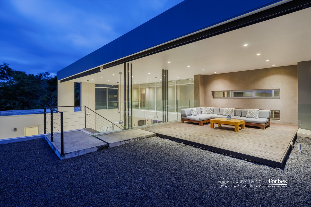 Modern home terrace with glass walls, a gray sectional, and a raised wood deck at dusk.