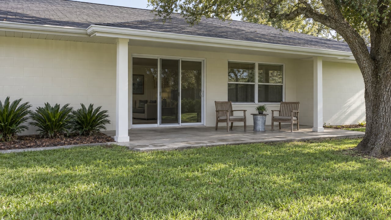 A modest off-white ranch with a shaded rear patio, two teak chairs, and a broad lawn beside a mature live oak.