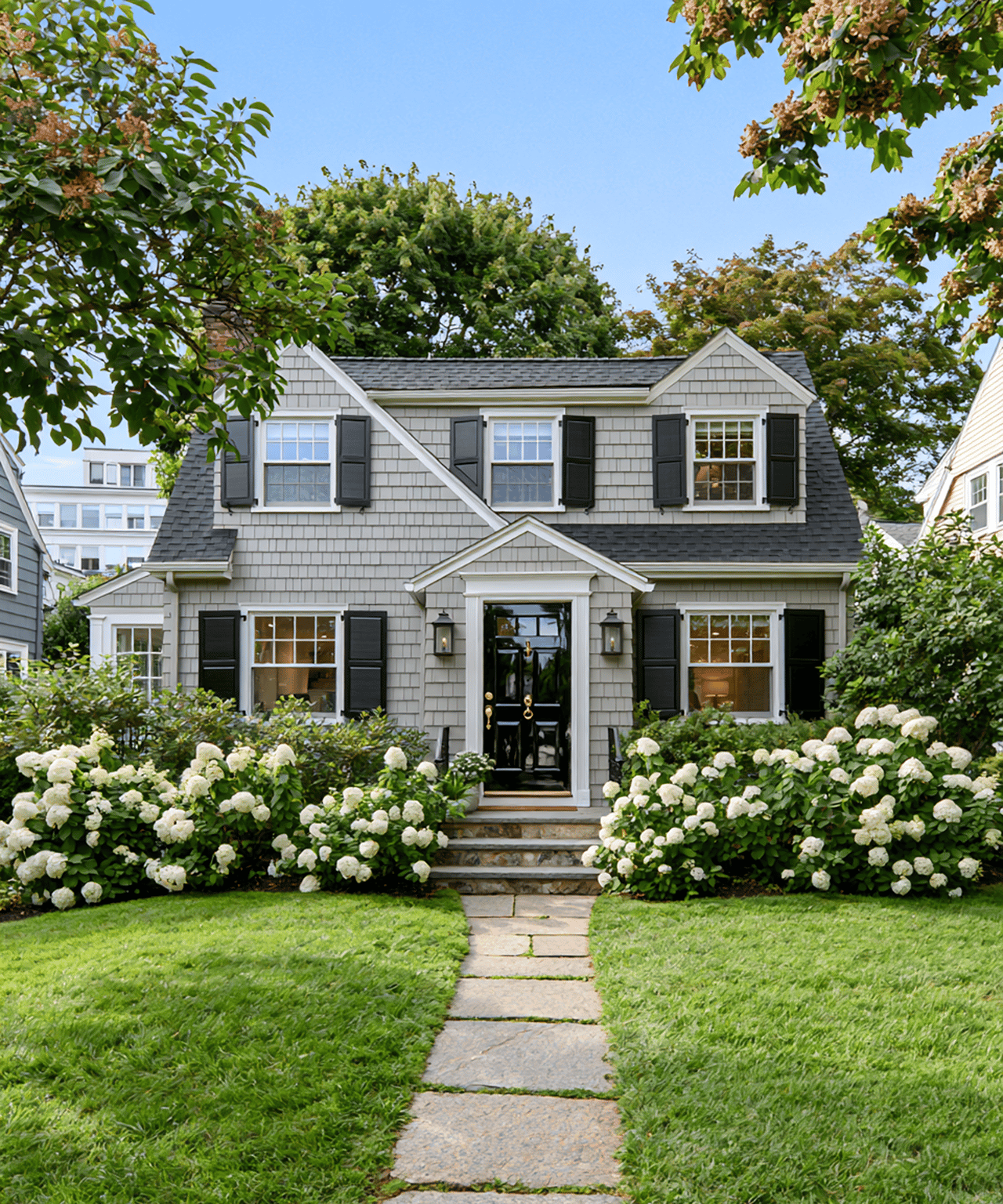 Gray shingle-sided house with black shutters, white hydrangeas, and a stone walkway to the front door.