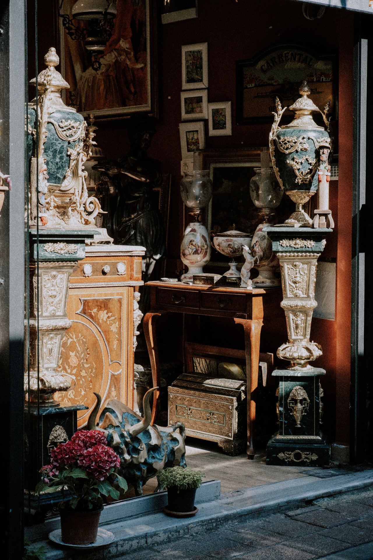 Antique shop window filled with ornate vases, carved furniture, figurines, and potted flowers.