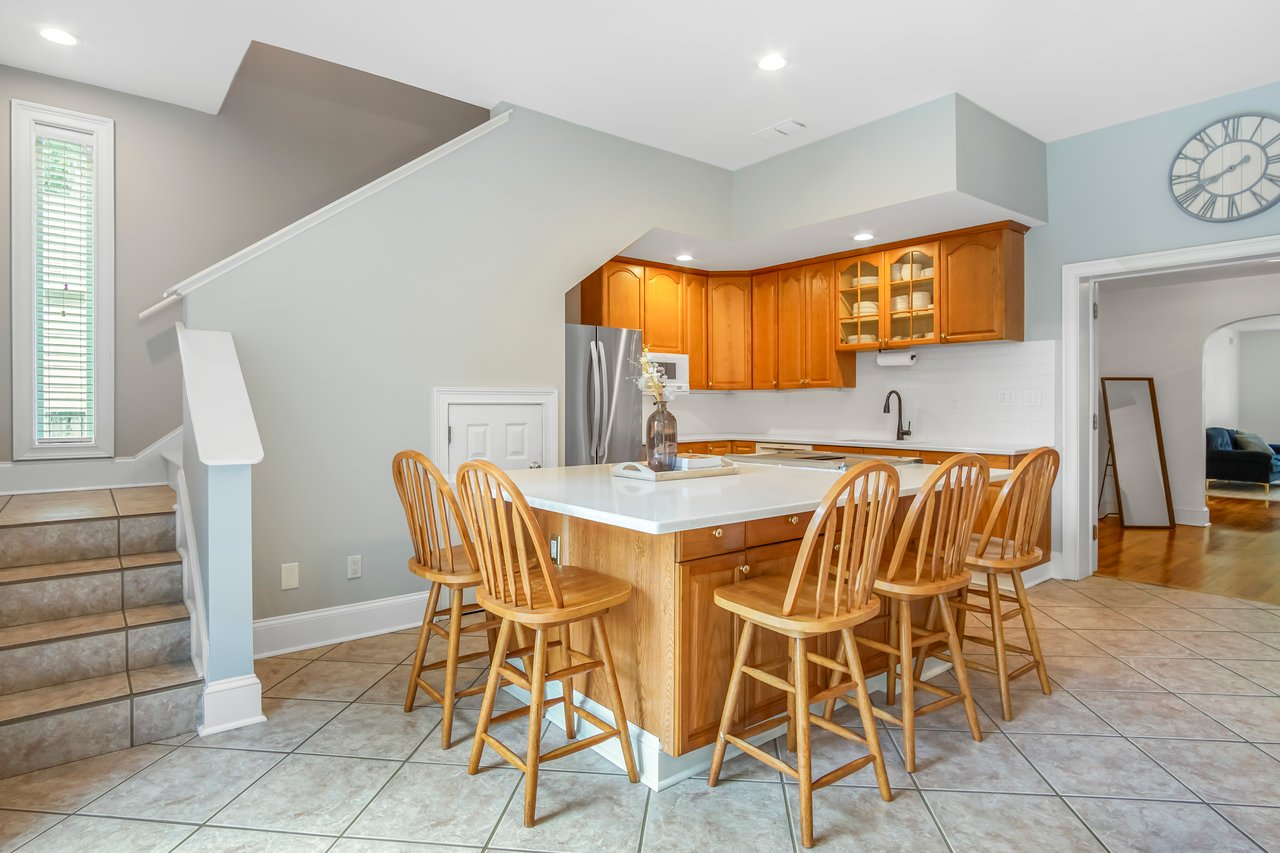 A cozy kitchen with light wood cabinets and a central island bordered by six wooden chairs. Warm lighting enhances the inviting, homely atmosphere.