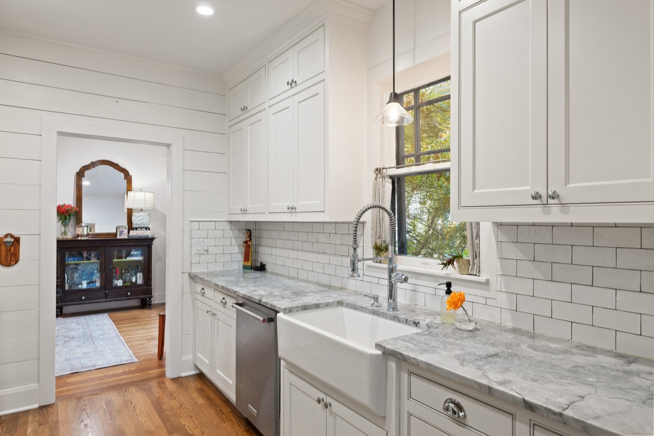 Bright white kitchen with a farmhouse sink, marble counters, subway tile, and a window overlooking trees.