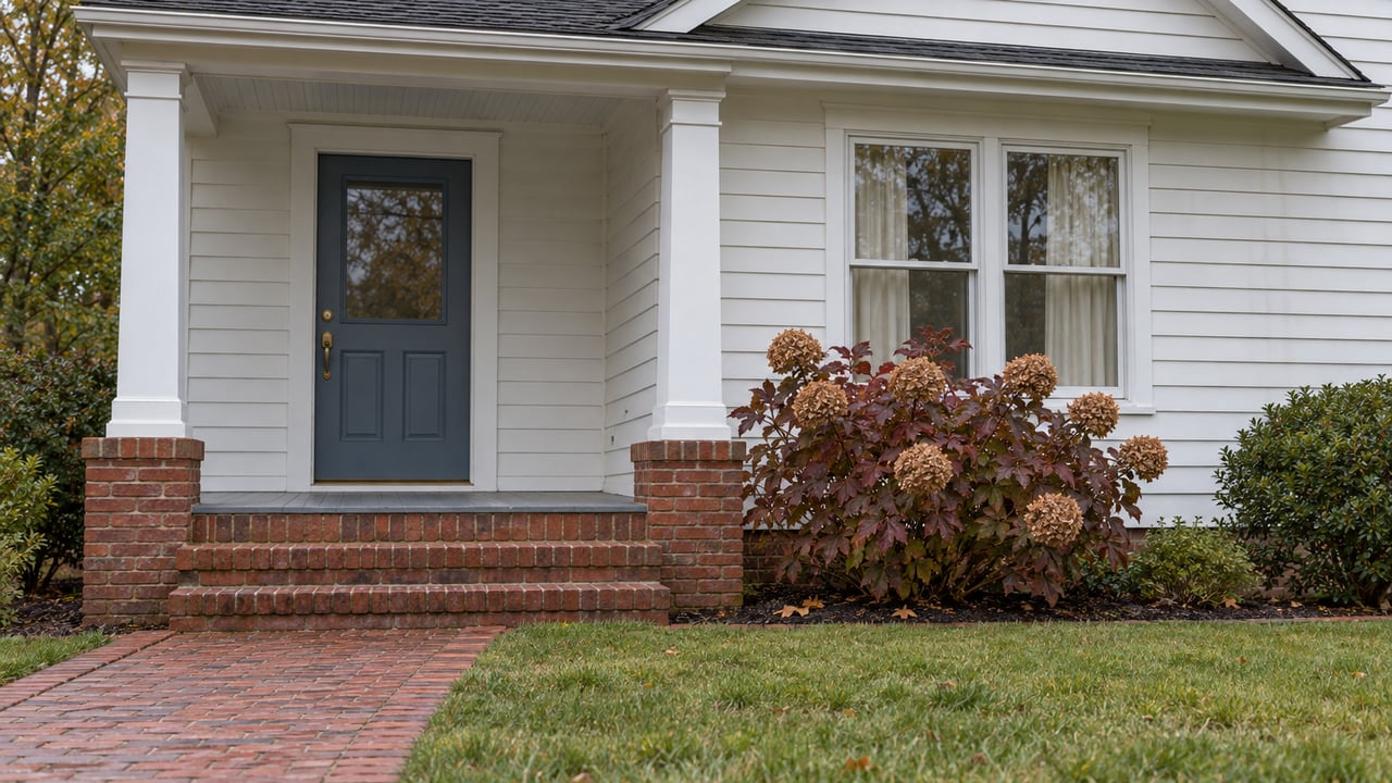 White clapboard home with a brick approach, gray timber porch, blue front door and burgundy oakleaf hydrangea in soft autumn daylight.