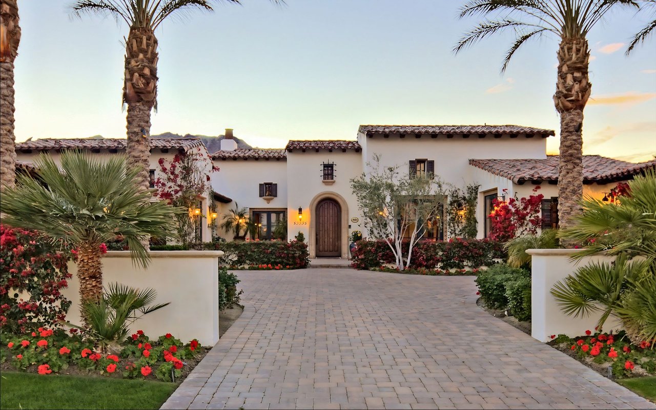 Spanish-style stucco home framed by palm trees, with a paved courtyard and flowering landscaping.