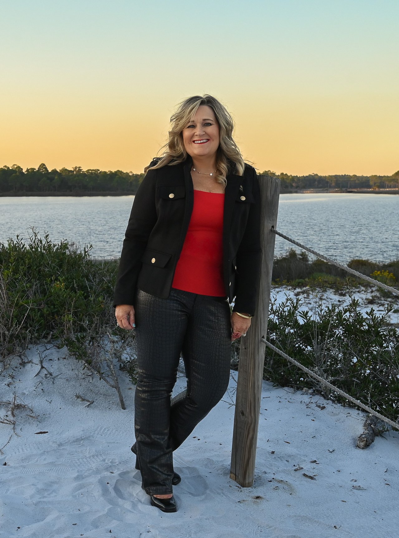 Smiling woman in a black jacket and red top beside a sandy waterfront at sunset.
