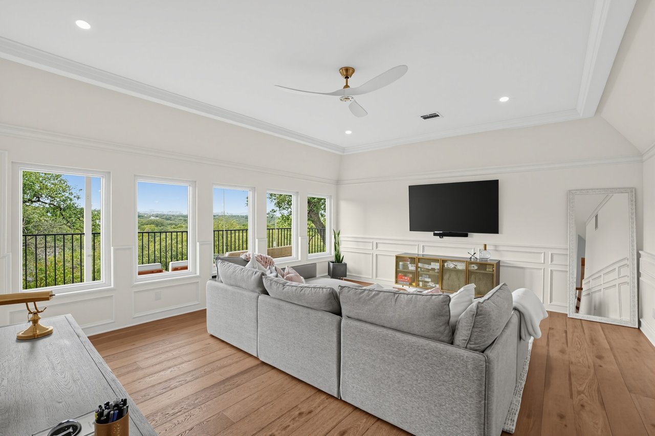 Living room with a gray sectional, white paneled walls, and tall windows overlooking leafy trees.