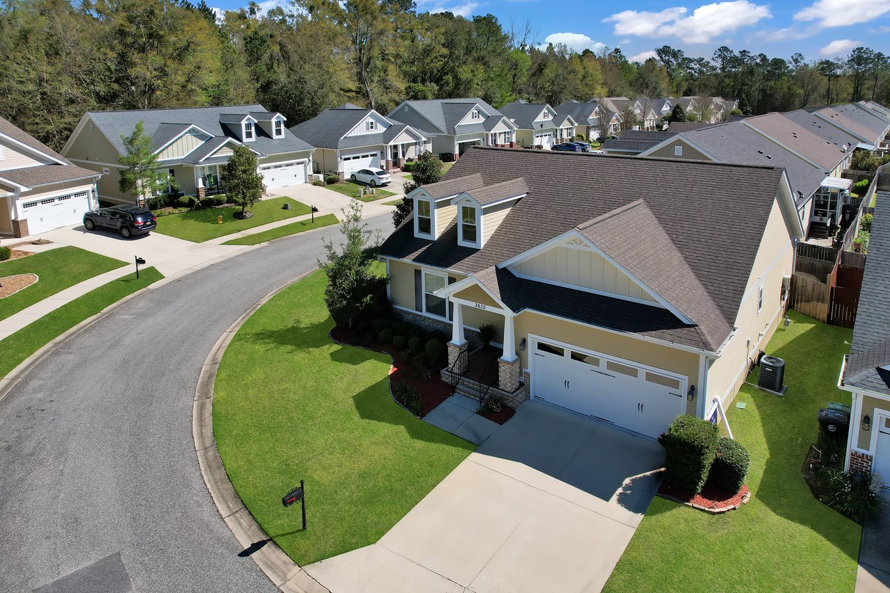 Aerial view of a suburban neighborhood with identical beige and gray houses, lined along a curved street. Neat lawns and green trees enhance the serene atmosphere.