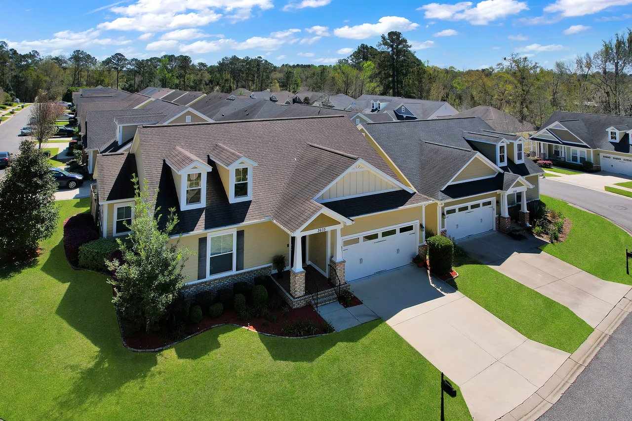 Aerial view of a suburban neighborhood with identical yellow houses. They have gabled roofs, two-car garages, and well-kept lawns on a sunny day.