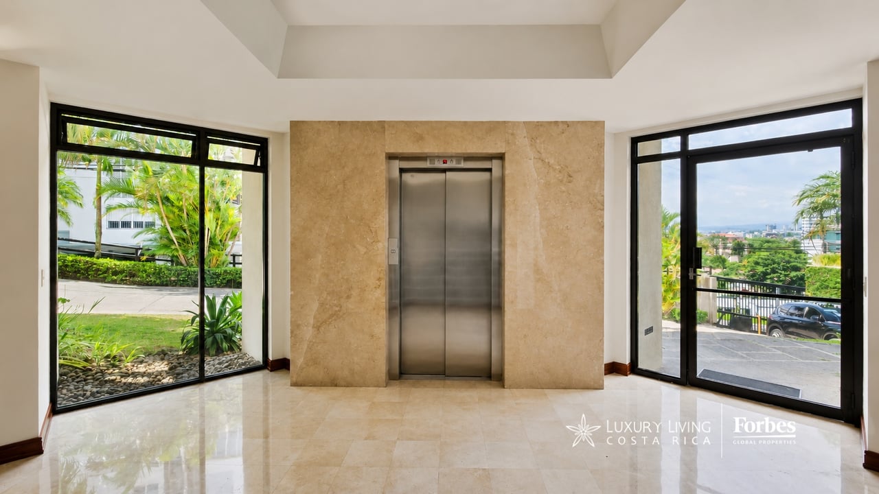 Condominium lobby with a central elevator, polished stone floor, and glass doors overlooking greenery.