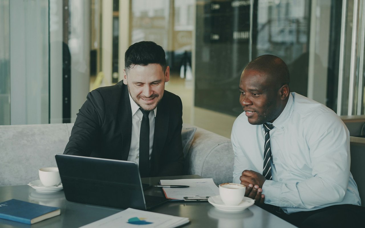 Two men review paperwork and a laptop together in a modern office lounge.