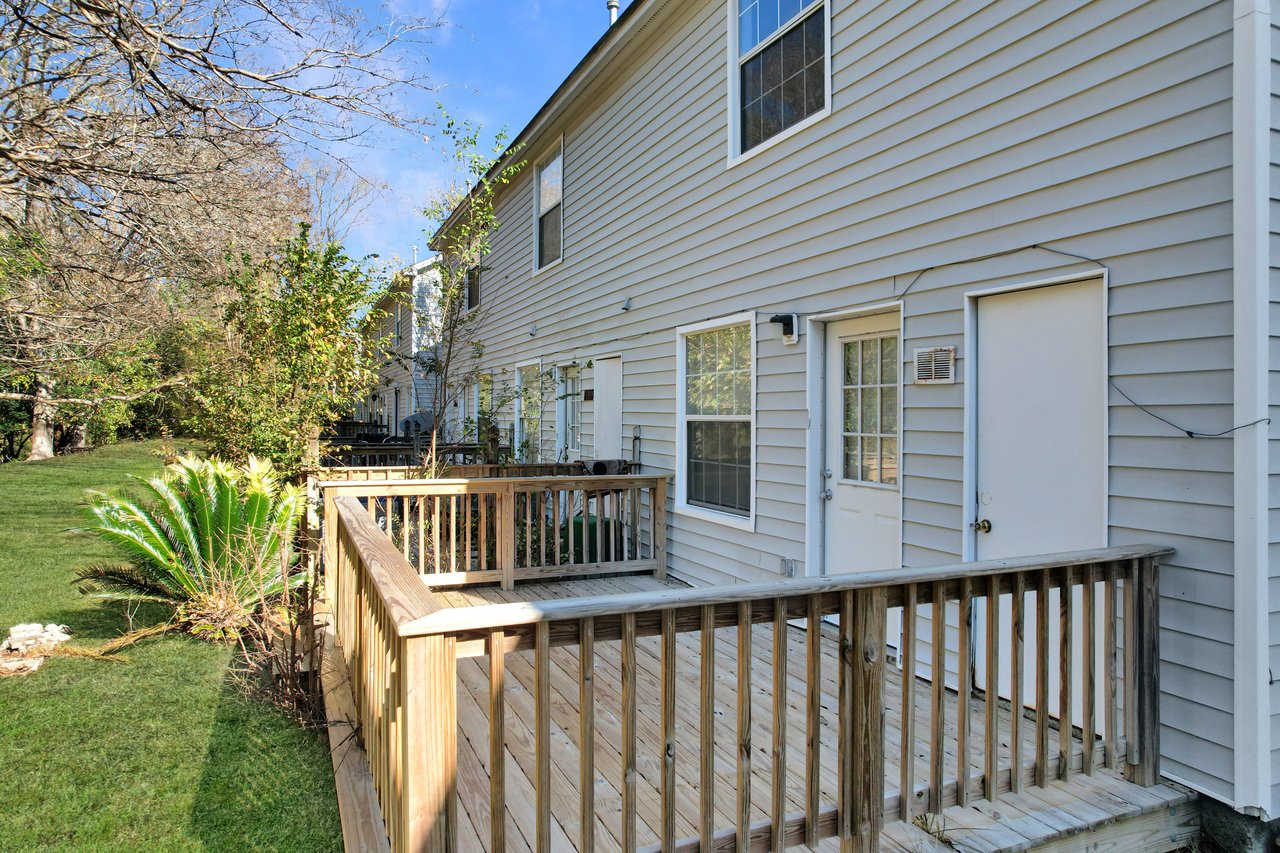 Wooden deck attached to the back of a grey house with a grassy yard and trees. White doors and windows are visible on the house exterior.