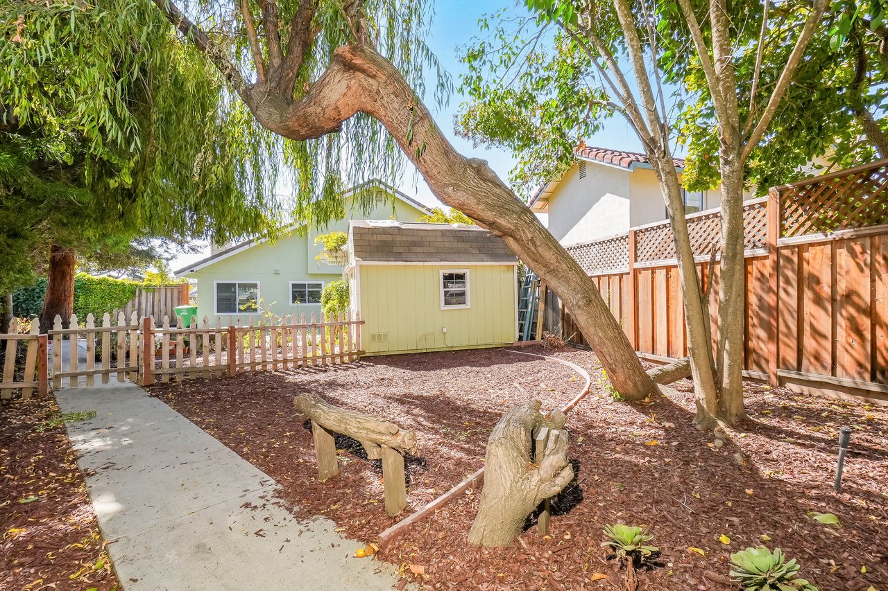 Backyard with a yellow shed, mature trees, mulch beds, and a concrete path.