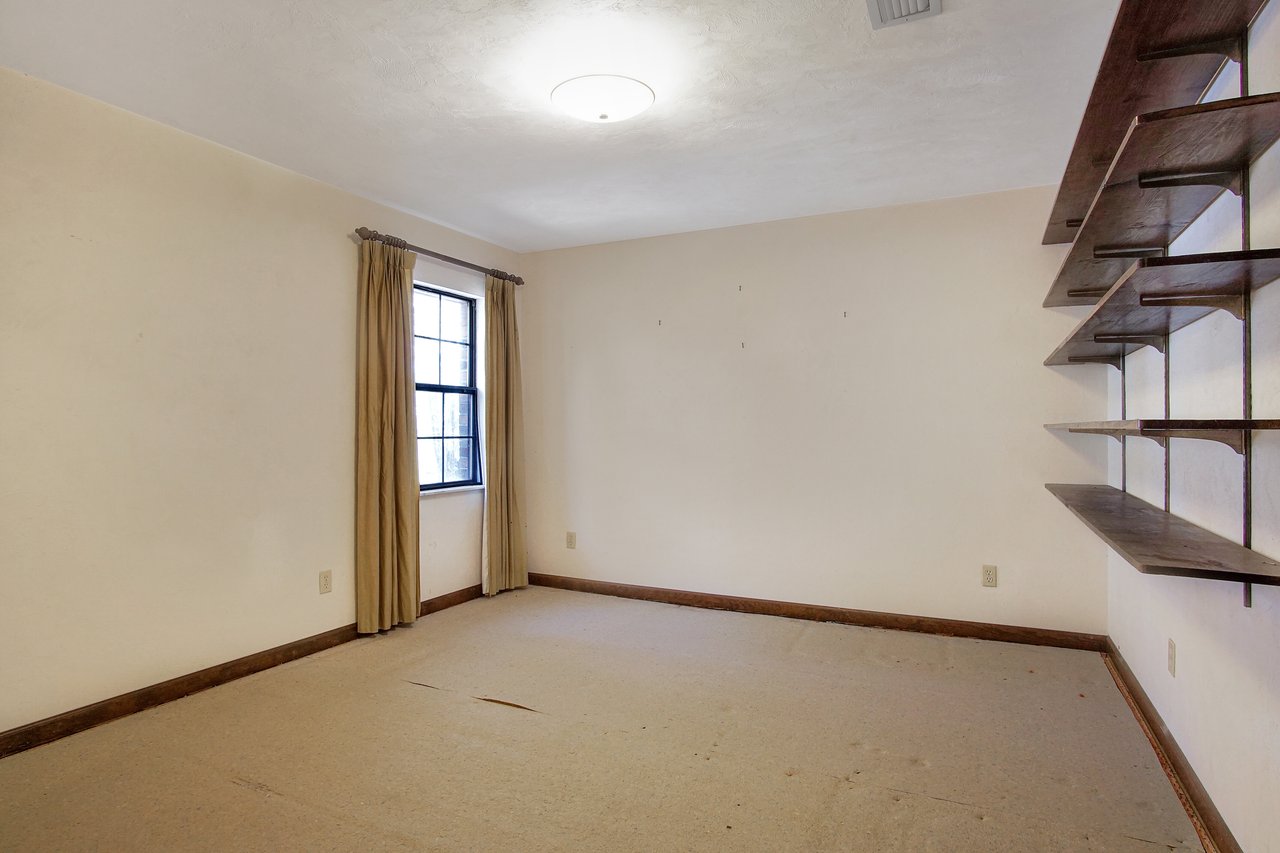 Empty room with beige walls and carpet, featuring a single window with tan curtains on the left. Wooden shelves on the right wall, under a ceiling light.