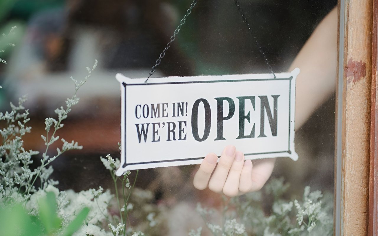 An open sign hangs in a storefront window, held from behind by a hand.