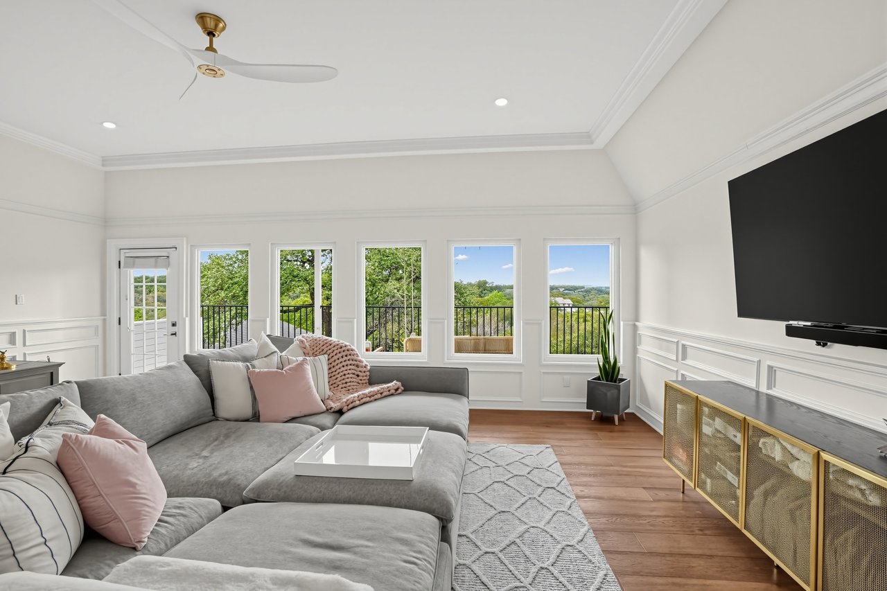 Living room with a gray sectional, wall-mounted TV, and tall windows overlooking trees.