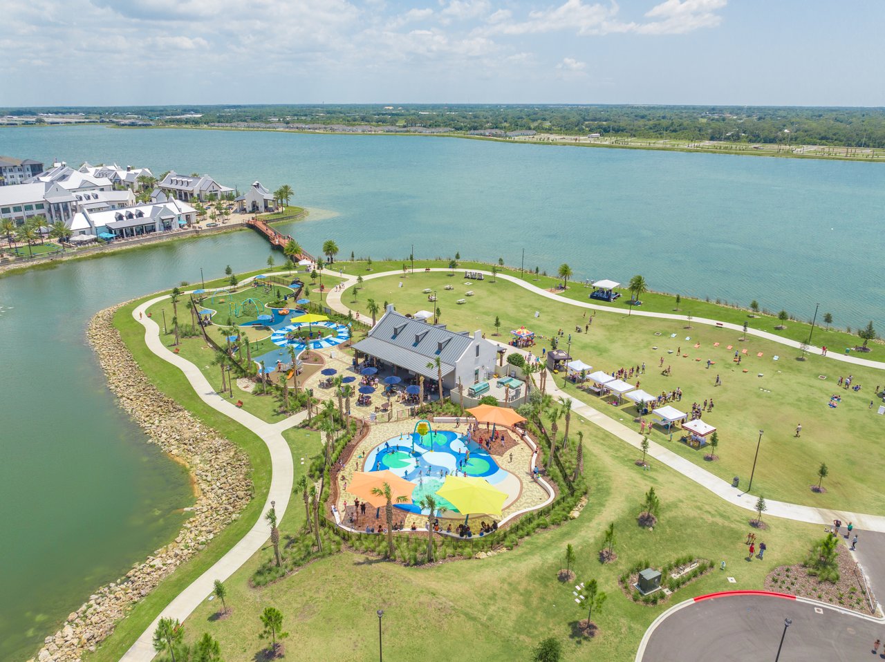 Aerial view of a waterfront park with a splash pad, playground, walking paths, and lakefront buildings.
