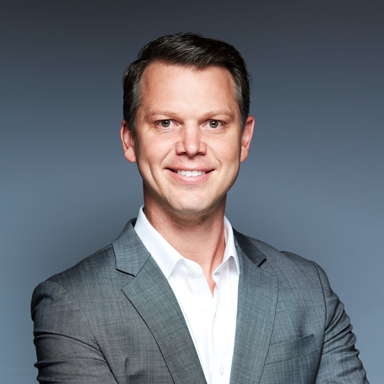 Smiling man in a gray suit jacket and white shirt against a blue-gray background.