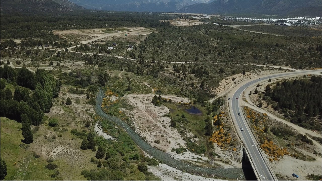 Aerial view of scrubland beside a river, with a highway bridge and distant mountains.