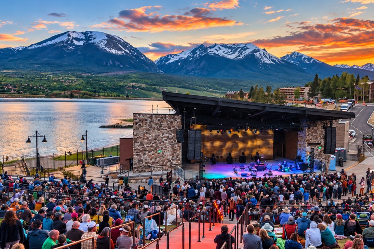 Outdoor concert at a lakeside amphitheater, backed by a lake and snow-dusted mountains at sunset.