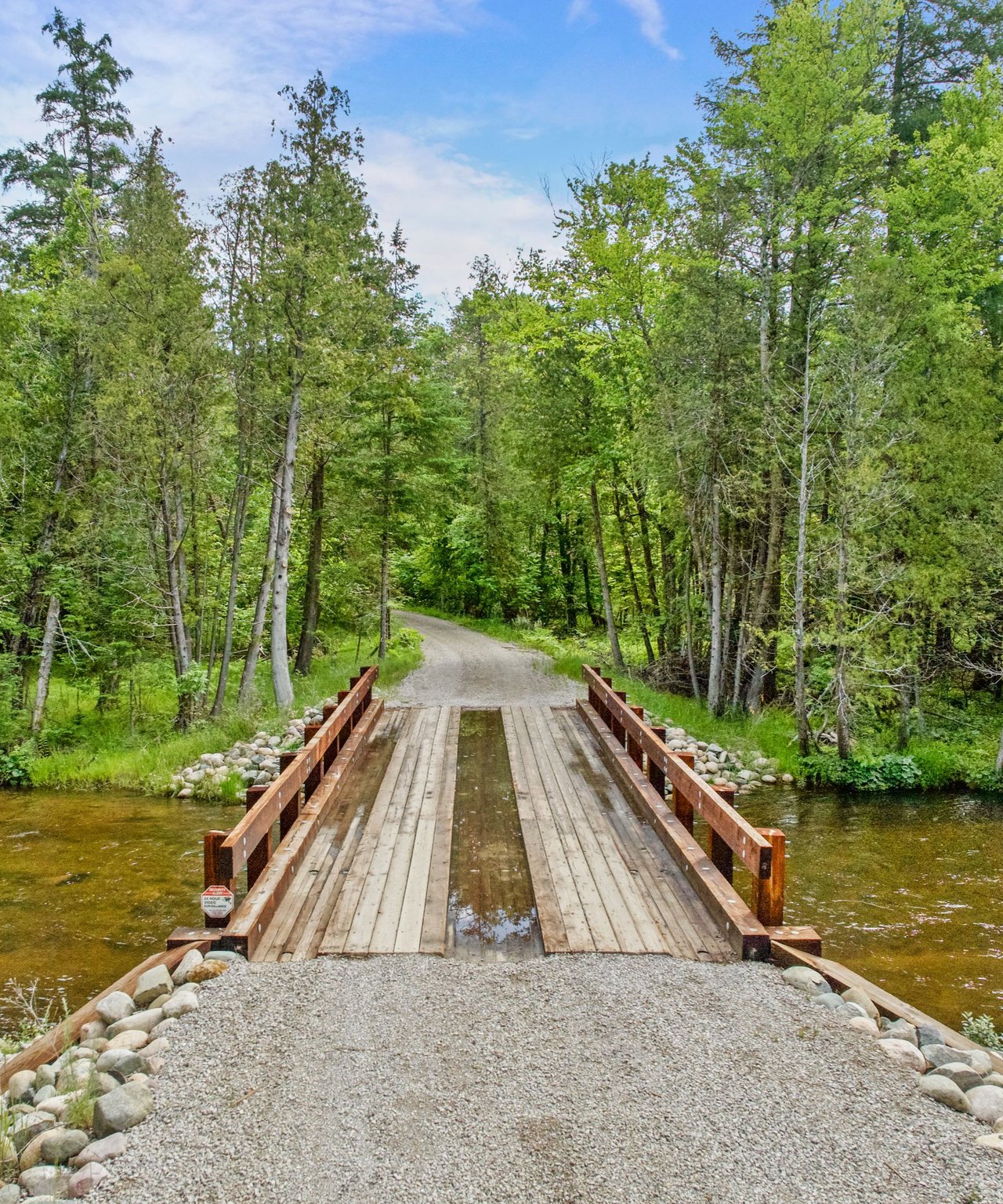 Wooden bridge crosses a shallow stream into a leafy woodland path.
