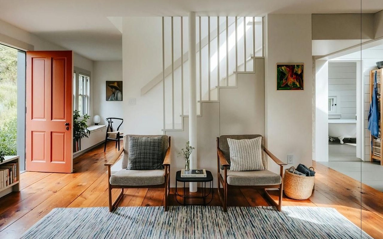 Living room with a coral-red front door, white staircase, and two chairs on hardwood floors.