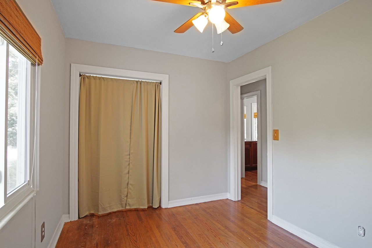 Bright room with wood floor, gray walls, and ceiling fan. A window with blinds on the left, a doorway with a curtain ahead, and a hallway on the right.