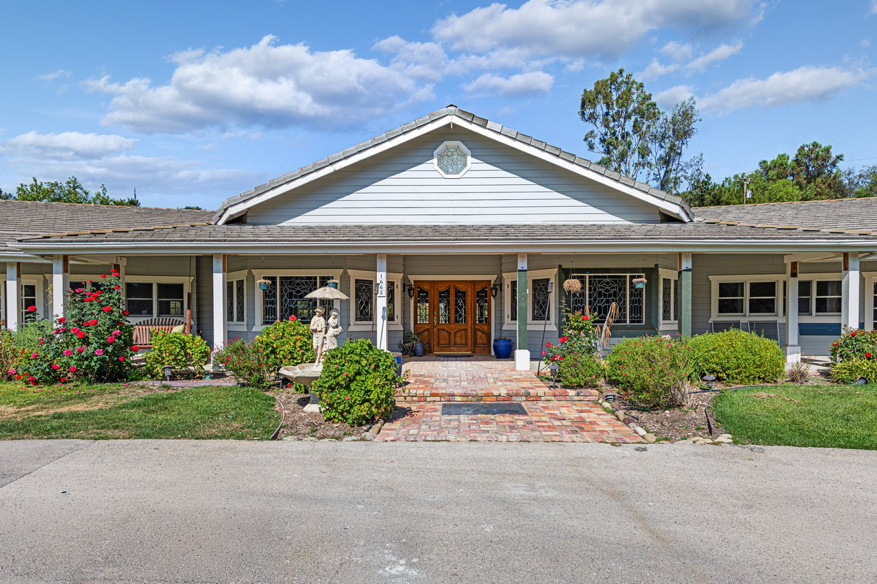 Gray single-story home with a covered porch, wooden double doors, and a brick walkway through flowering gardens.