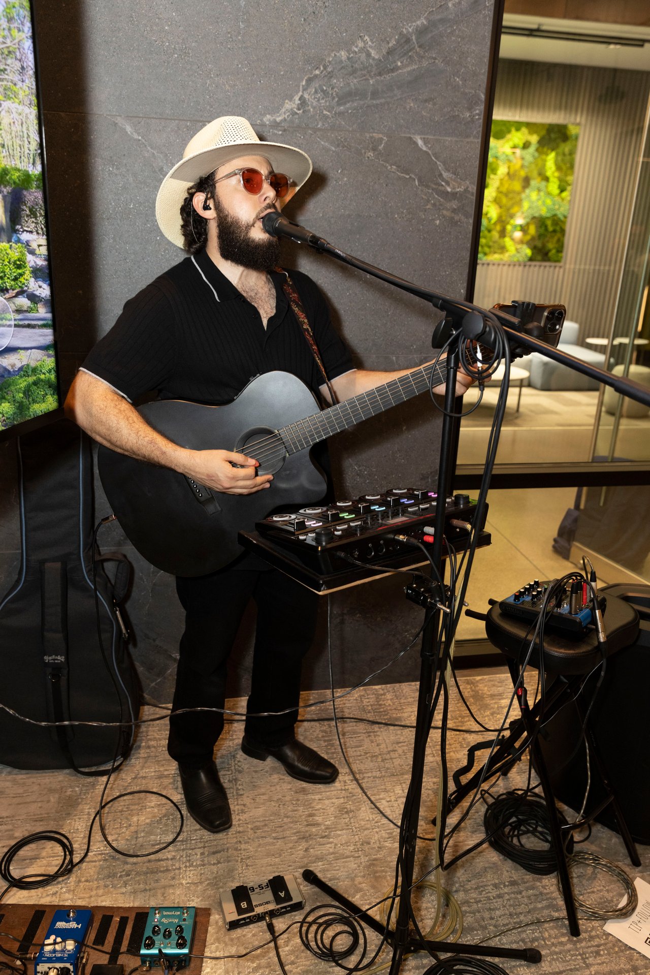 Bearded musician in a white hat performs on acoustic guitar with a microphone and effects pedals.