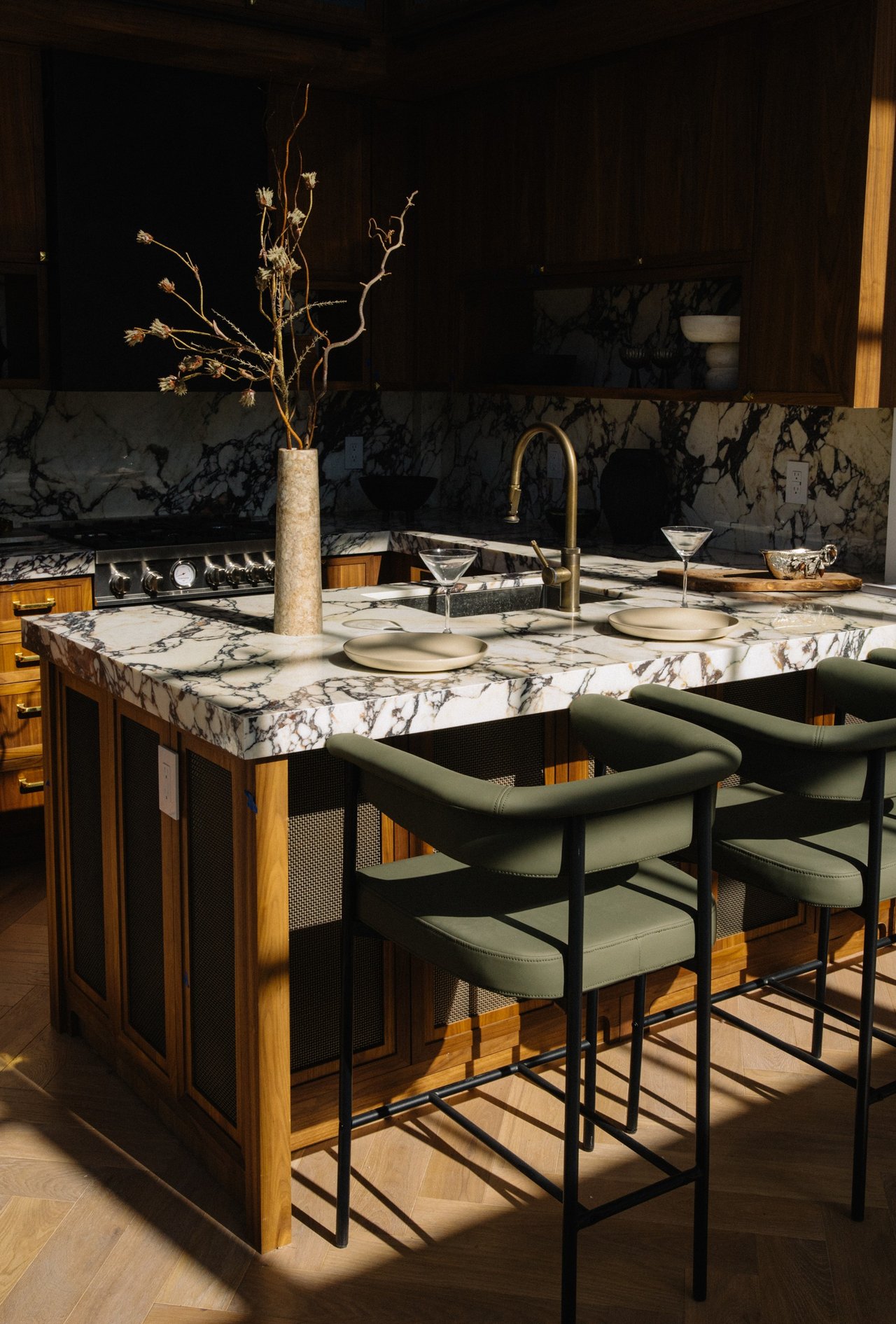 Kitchen with a marble island, dark green bar stools, wood cabinetry, and a brass faucet.