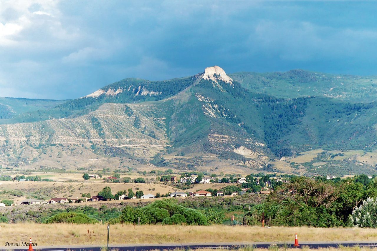 Battlement Mesa homes below a forested mountain ridge with a pale rock outcrop.
