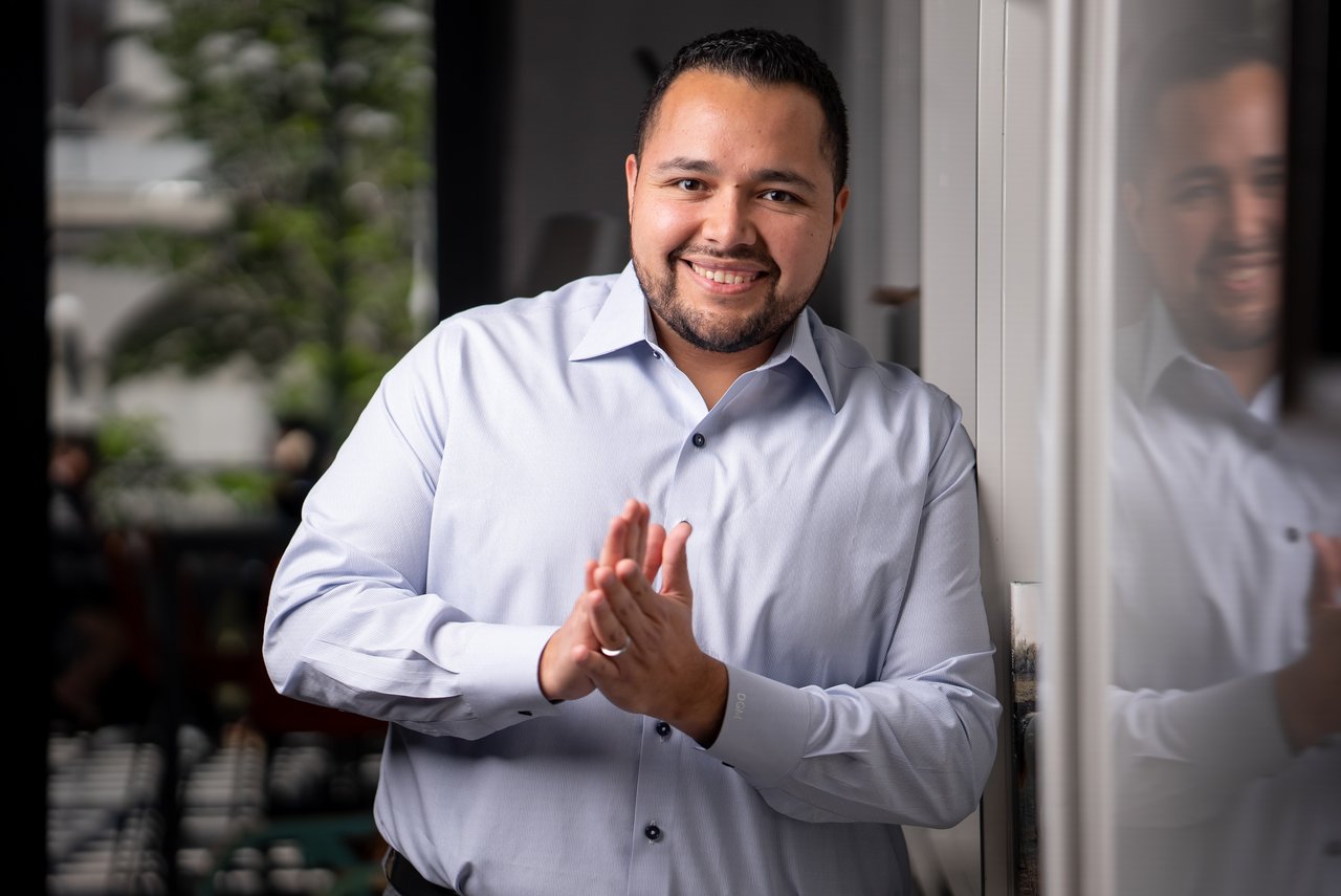 Smiling man in a light shirt standing beside a glass door, hands clasped.