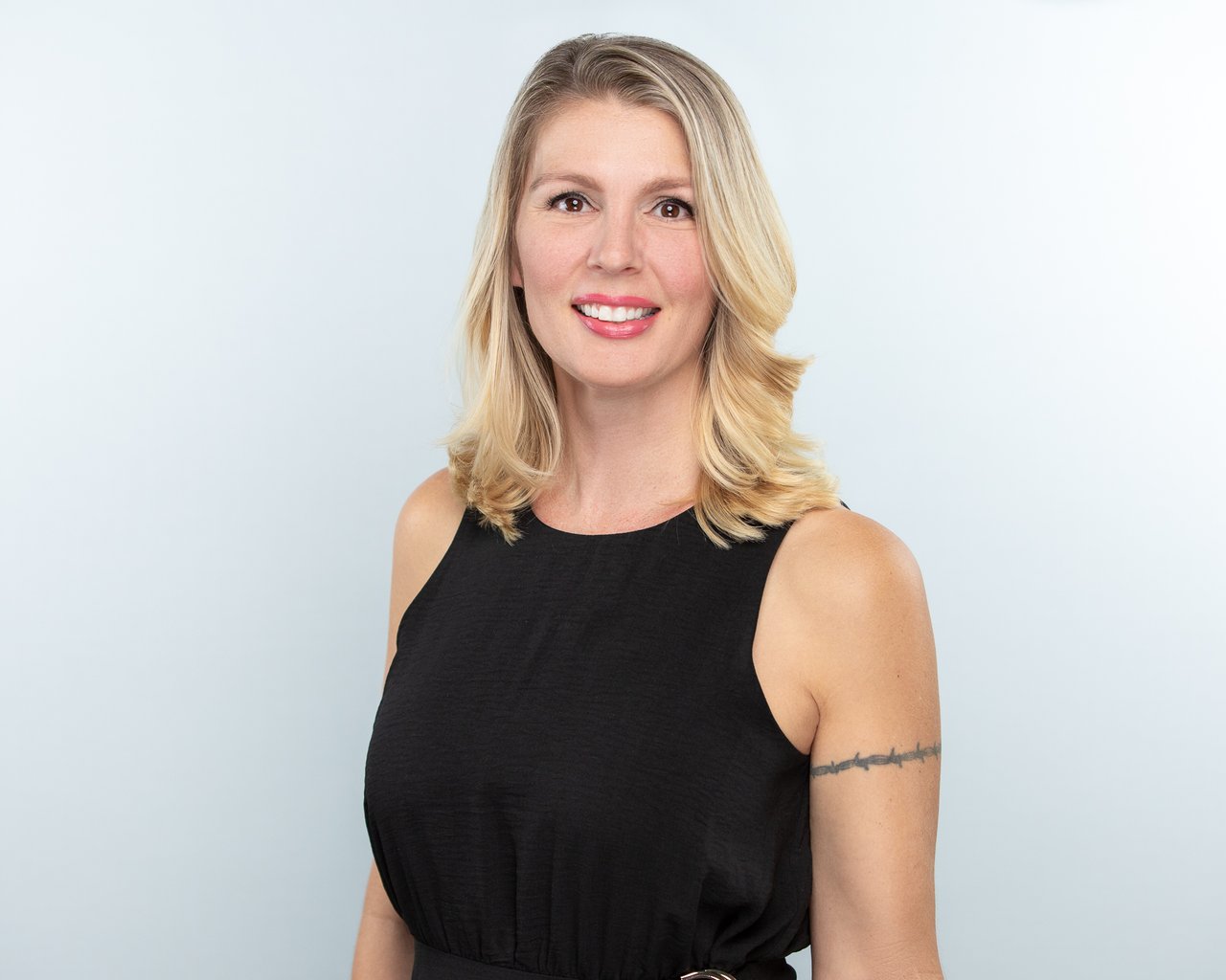 Smiling blonde woman in a black sleeveless top, with a tattoo on her upper arm.
