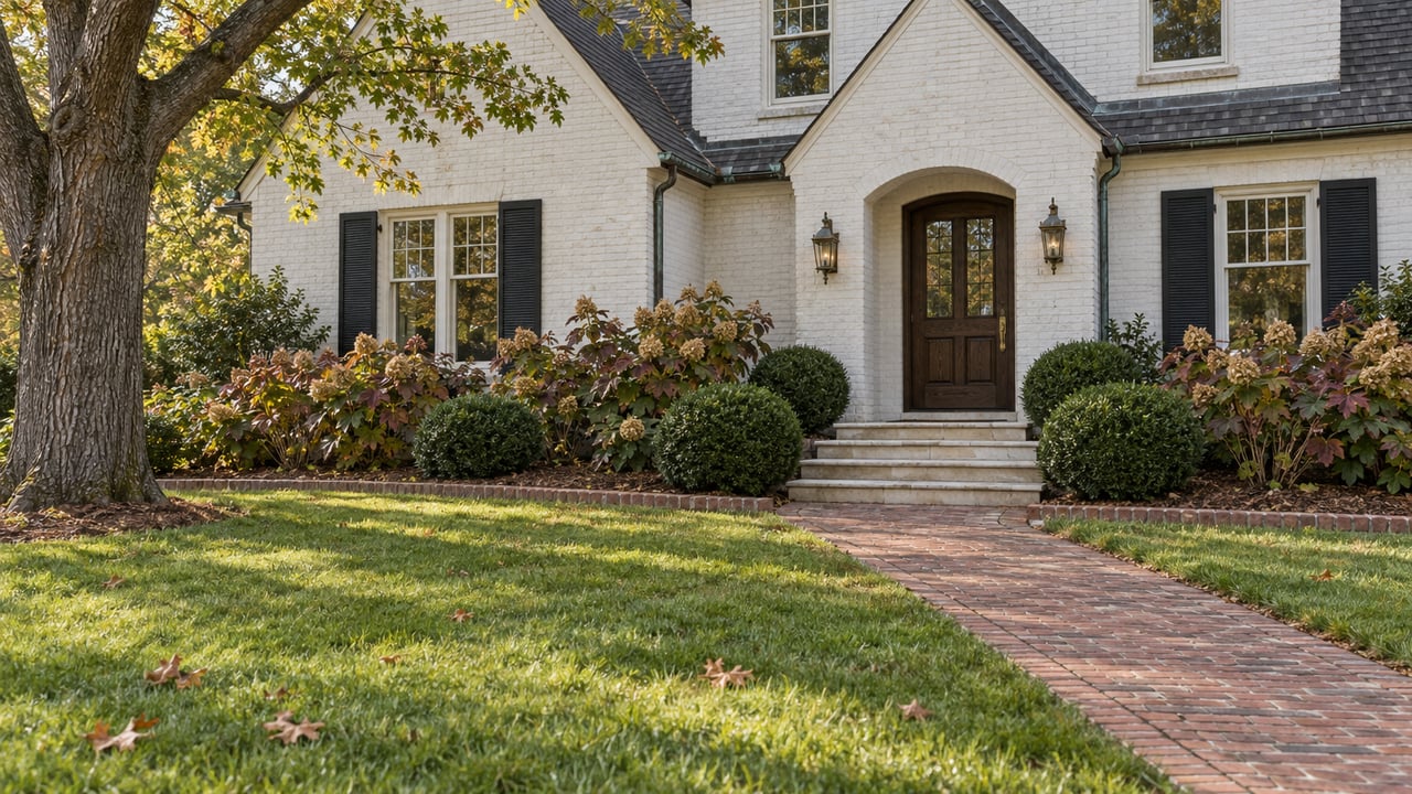White-painted brick home with black shutters, an arched dark-wood entry, and a brick walk through an autumn front garden.