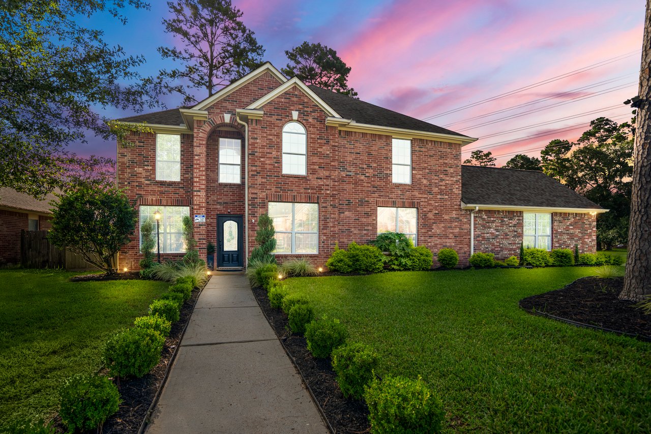 Two-story brick house with a front walkway, landscaped lawn, and pink dusk sky.