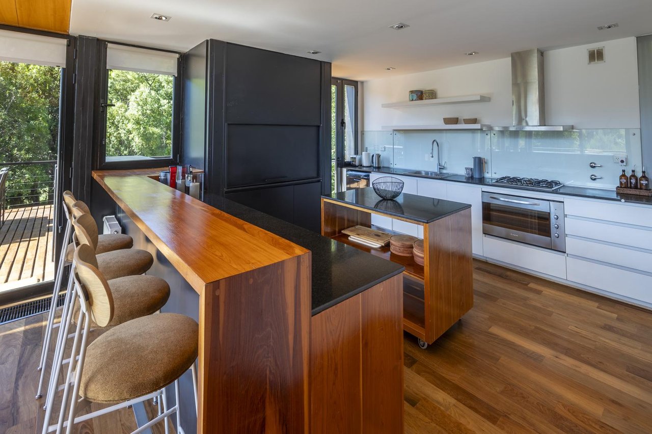 Modern kitchen with a long wooden breakfast bar, stools, and large windows overlooking trees.