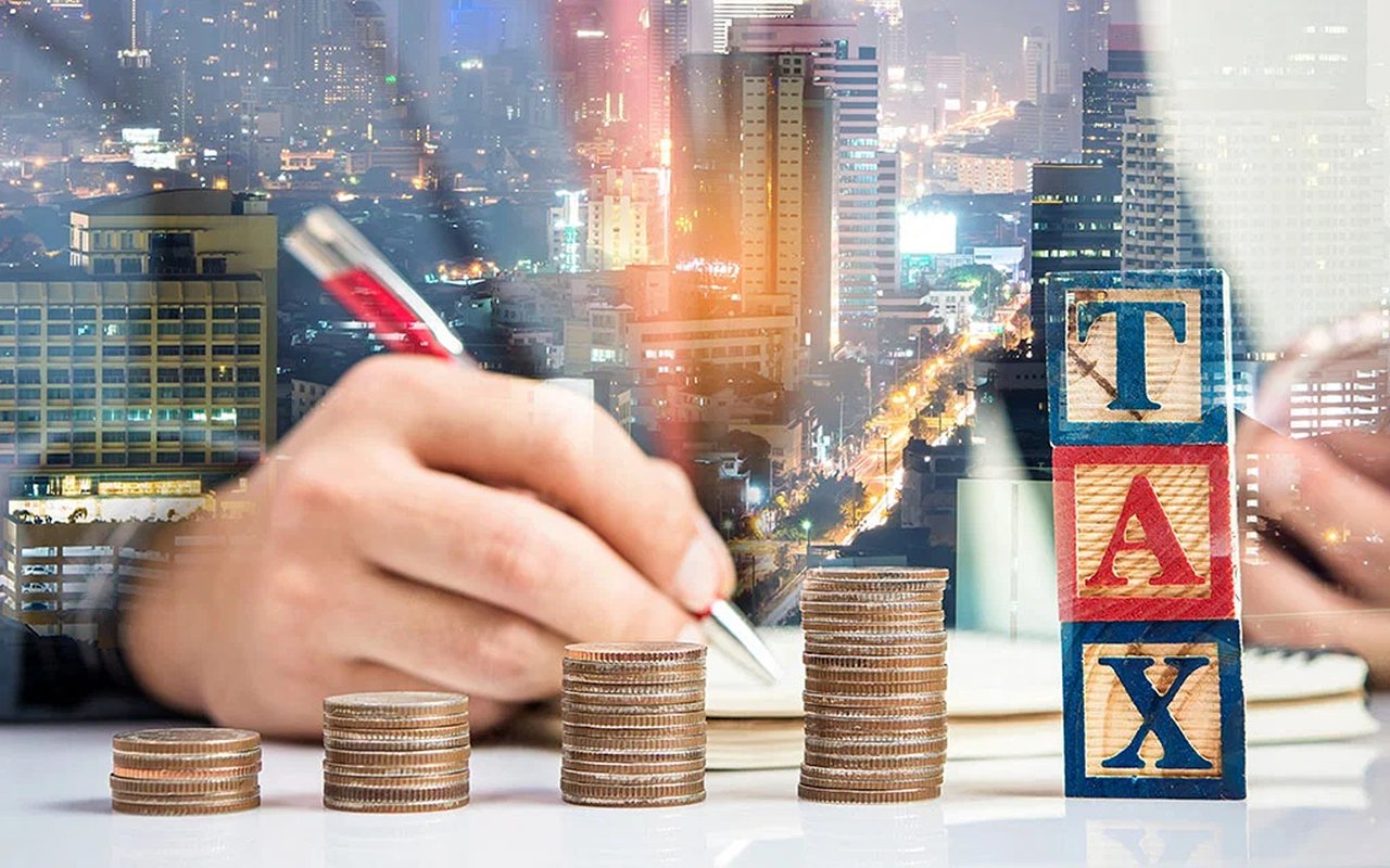 Tax blocks and rising coin stacks beside a hand writing against a nighttime city skyline.