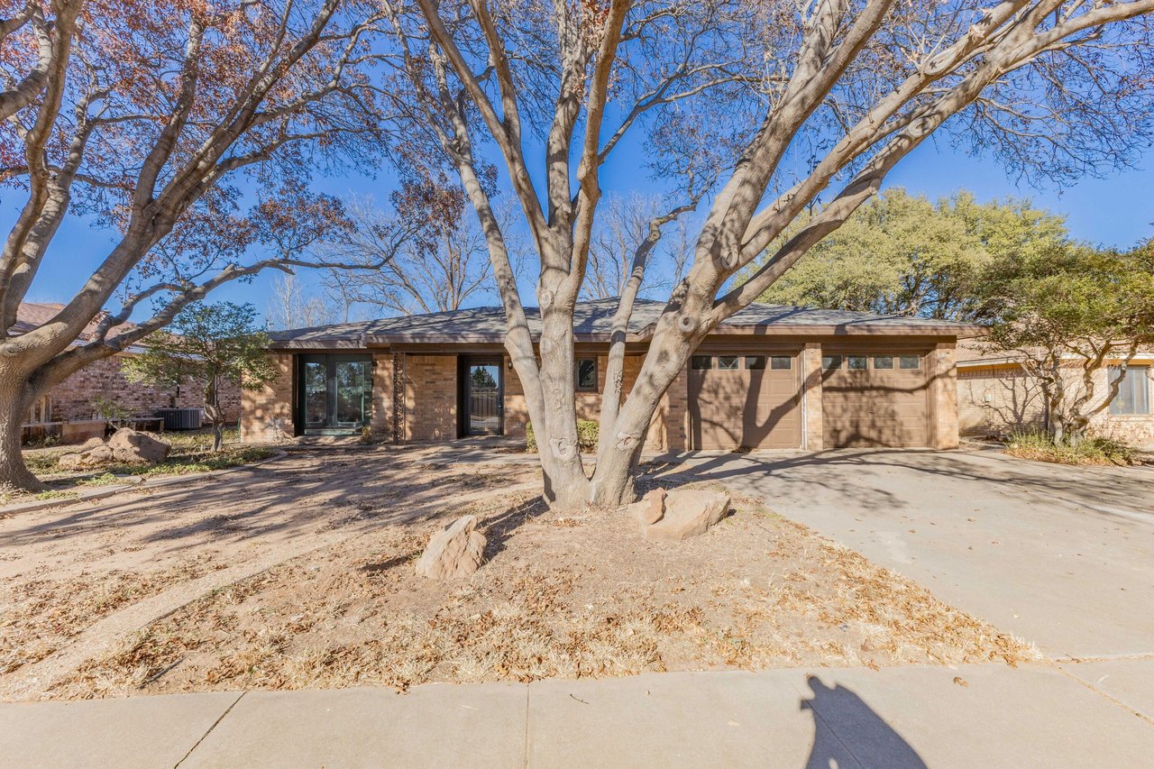 Single-story brick home with an attached garage, framed by large mature trees.