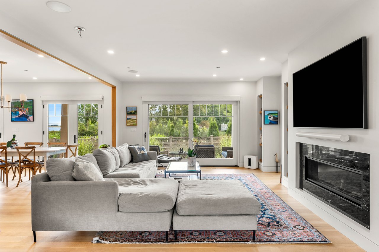 Living room with a gray sectional, fireplace, and broad windows overlooking a green yard and water.
