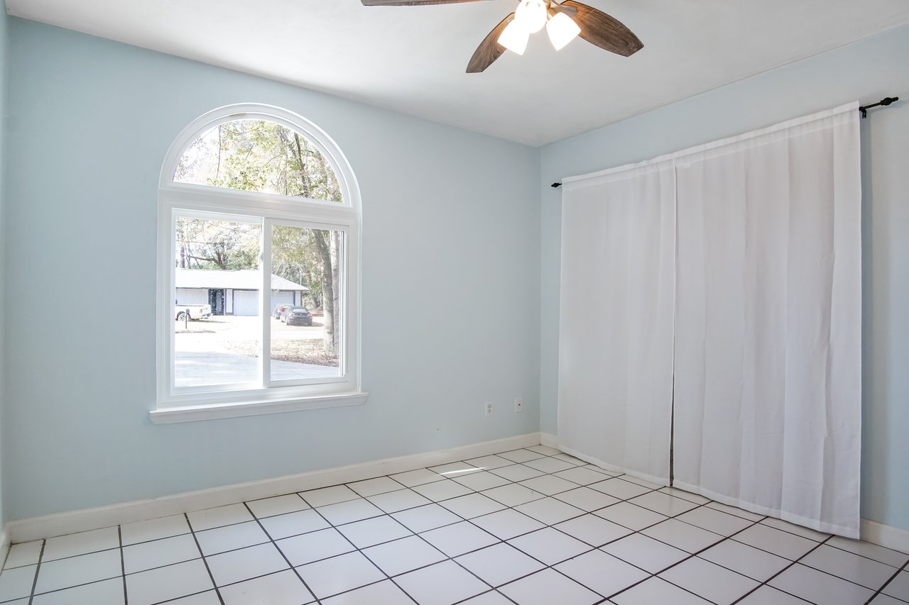 A bright, empty room with light blue walls, a large arched window showing trees, white tiled floor, ceiling fan, and white curtains. Relaxing atmosphere.