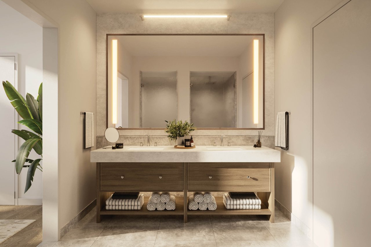 Bathroom with a double-sink wood vanity, illuminated mirror, folded towels, and a leafy plant.