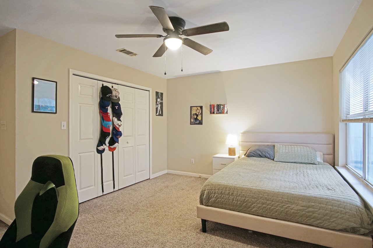 Bedroom at 529 Collinsford Road, Tallahassee, Florida, featuring beige walls, carpet flooring, and a ceiling fan with light fixture. The room includes a bed with green bedding, nightstand with lamp, large window with blinds, and a white double-door closet. Wall art and hats add a personal touch to the space.