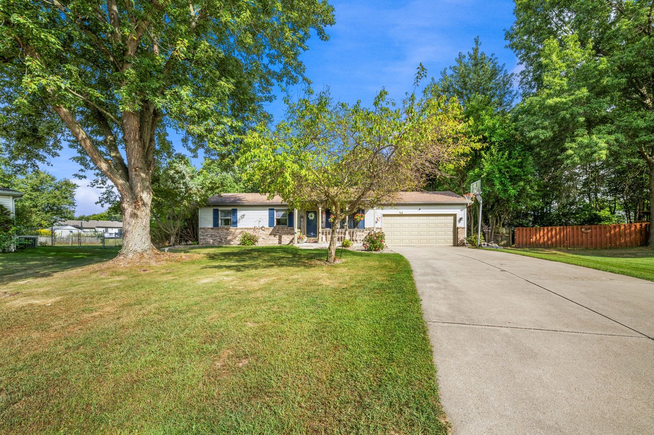 Single-story home with stone façade, attached garage, broad driveway, and mature shade trees.